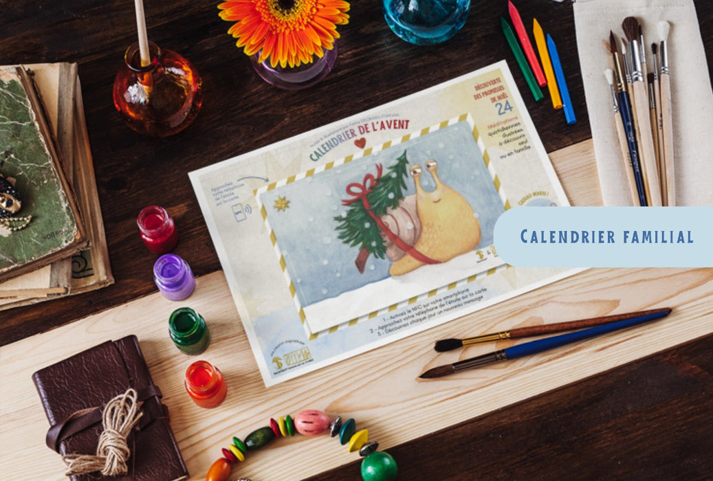 Le calendrier de l'avent est posé sur un bureau de travail. Su ce calendrier de l'avent il y a l'escargot qui porte sur sa coquille un sapin de Noël. Autour du calendrier de l'avant il y a de la peinture , des pinceaux et des crayons.
