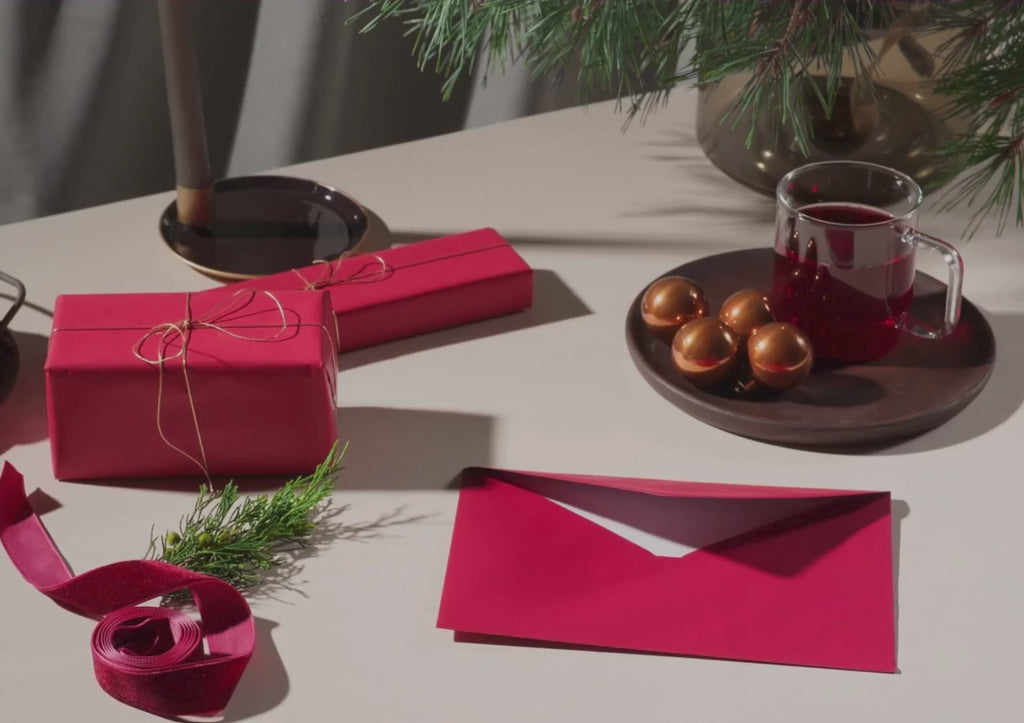 Une table est dressée pour Noël, la nappe est blanche il y a deux cadeaux avec du papier cadeau rouge et une enveloppe semi-ouverte rouge. Il y a une tasse de thé posée sur une assiette et des boules de Noël dorées. Des mains tenant le calendrier de l'avent apparaissent sur le dessus de la table.